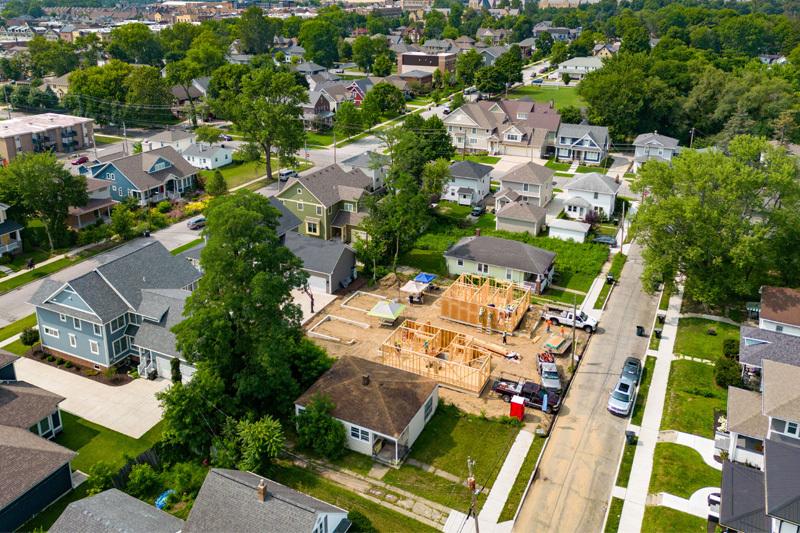 Aerial view of a residential neighborhood with two homes under construction. The wooden frames of the houses are visible, and construction workers are on site. Trucks and equipment are parked nearby. Surrounding the construction site are established homes with yards and driveways.